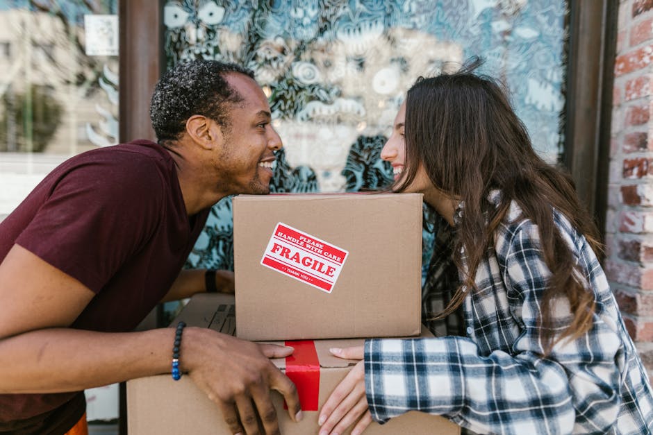 A man and a woman are outdoors, smiling and leaning close to each other across a stack of cardboard moving boxes. The man, with short curly hair and wearing a maroon t-shirt, is holding the boxes, whi