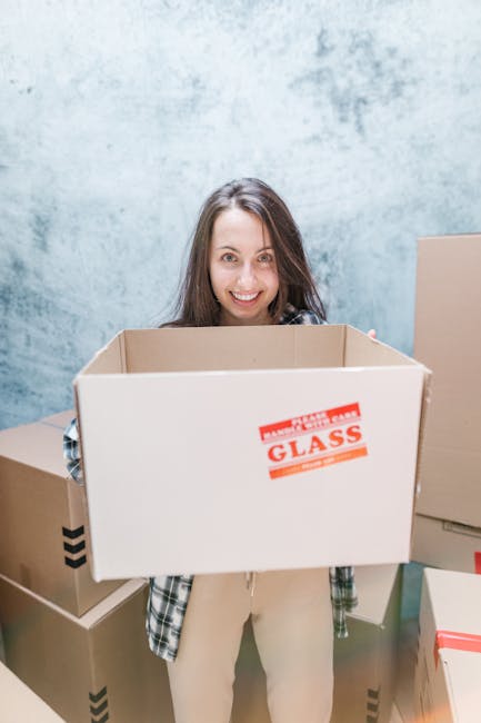 A man and a woman are outdoors, smiling and leaning close to each other across a stack of cardboard moving boxes. The man, with short curly hair and wearing a maroon t-shirt, is holding the boxes, whi